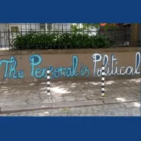 You can see a street wall with black railings and bushes coming through the railings. On the wall is some street art in blue and white which reads 'The Personal is Political'. 
