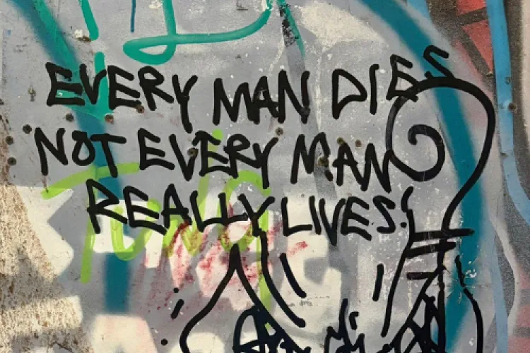 A heavily graffitied wall can be seen. Colours in the background are grey, with blue lines and faded white letters, in the foreground on the wall in black spray paint are the words: EVERY MAN DIES NOT EVERY MAN REALLY LIVES. 