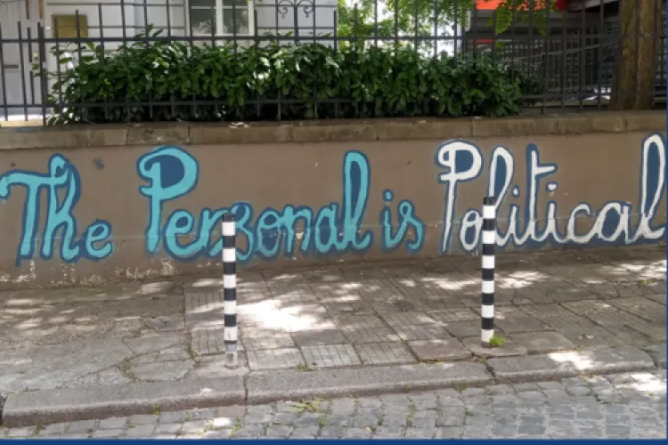 You can see a street wall with black railings and bushes coming through the railings. On the wall is some street art in blue and white which reads 'The Personal is Political'. 